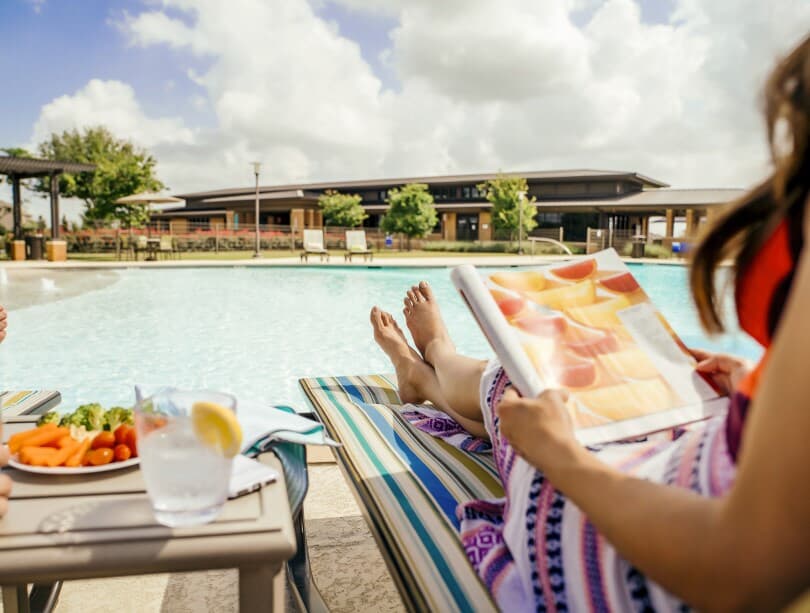 Woman lounging poolside with snacks and a magazine at Elyson by Brookfield Residential in Katy TX