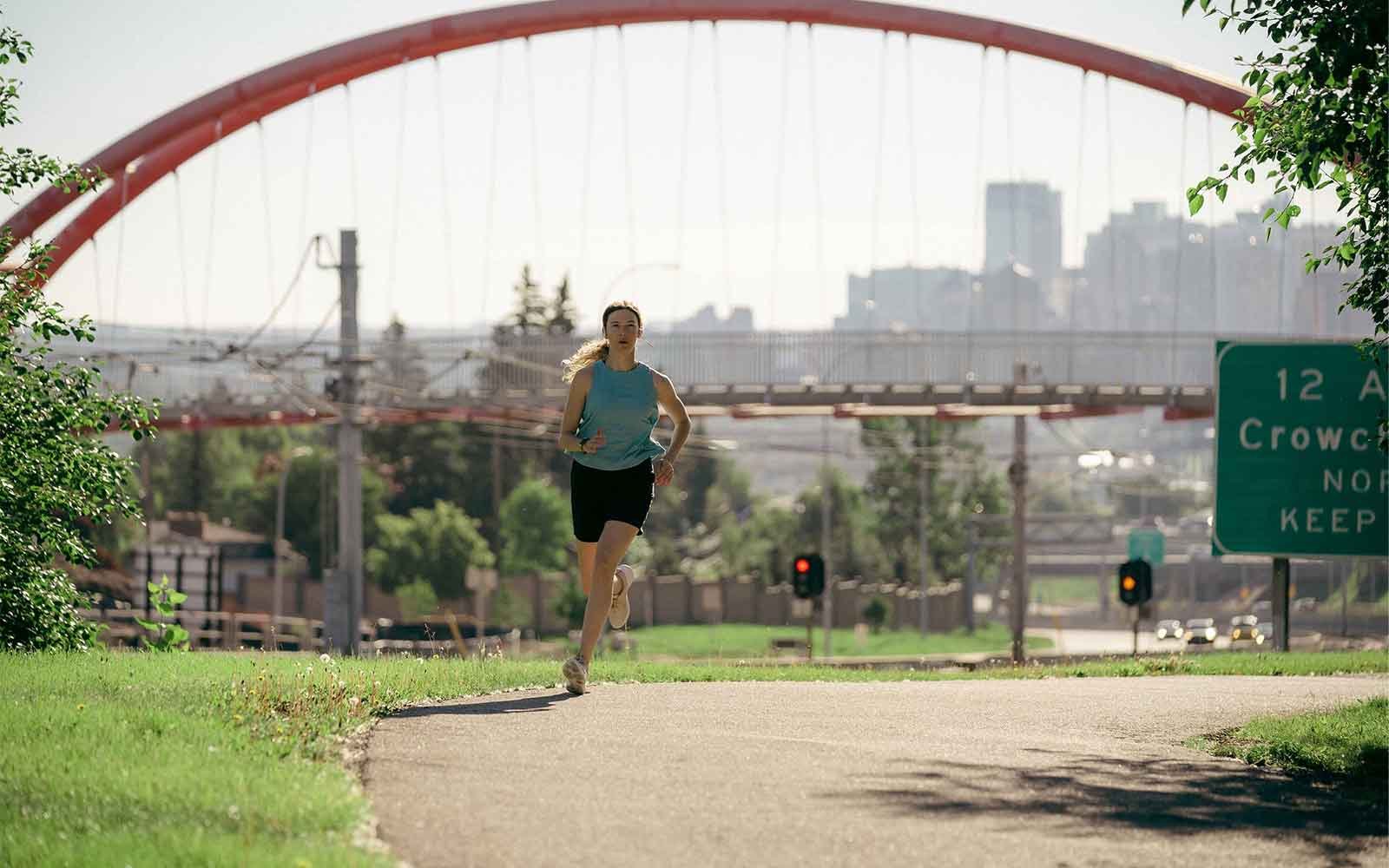 Woman running on a path near Crown Park with the pedestrian bridge and downtown Calgary in the background