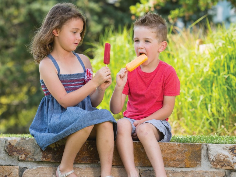 A boy and a girl sit on a wall eating popsicles 