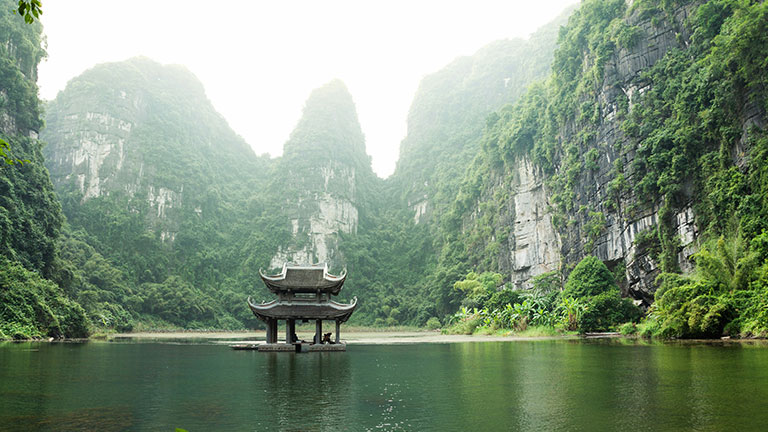 A traditional pavilion sits on a peaceful lake surrounded by towering limestone cliffs in Tràng An, Ninh Bình, Vietnam.