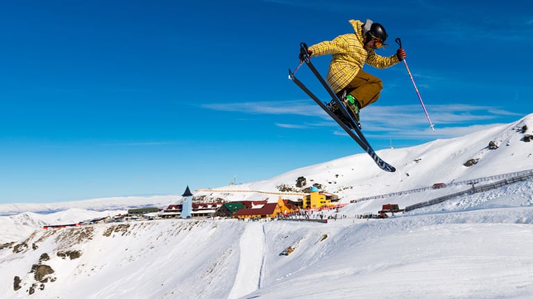A freestyle skier doing tricks in front of the Cardrona Mountain Ski Resort with plenty of snow and blue skies and visitors. Credit: CUHRIG