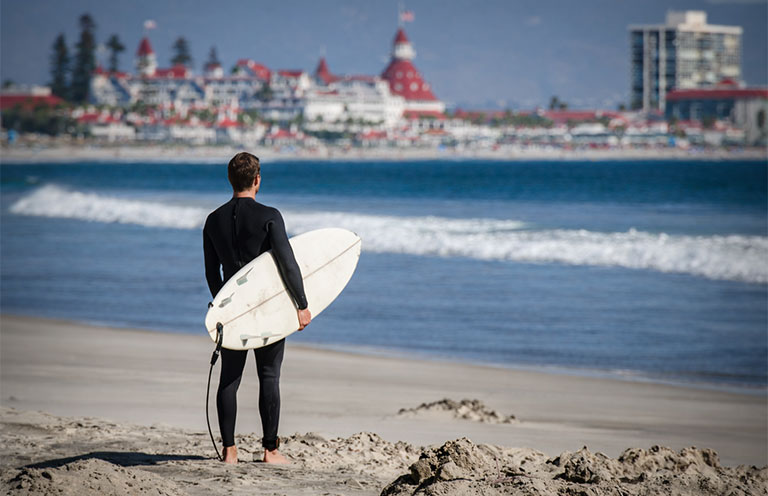man holding surf board Hotel del Coronado in background