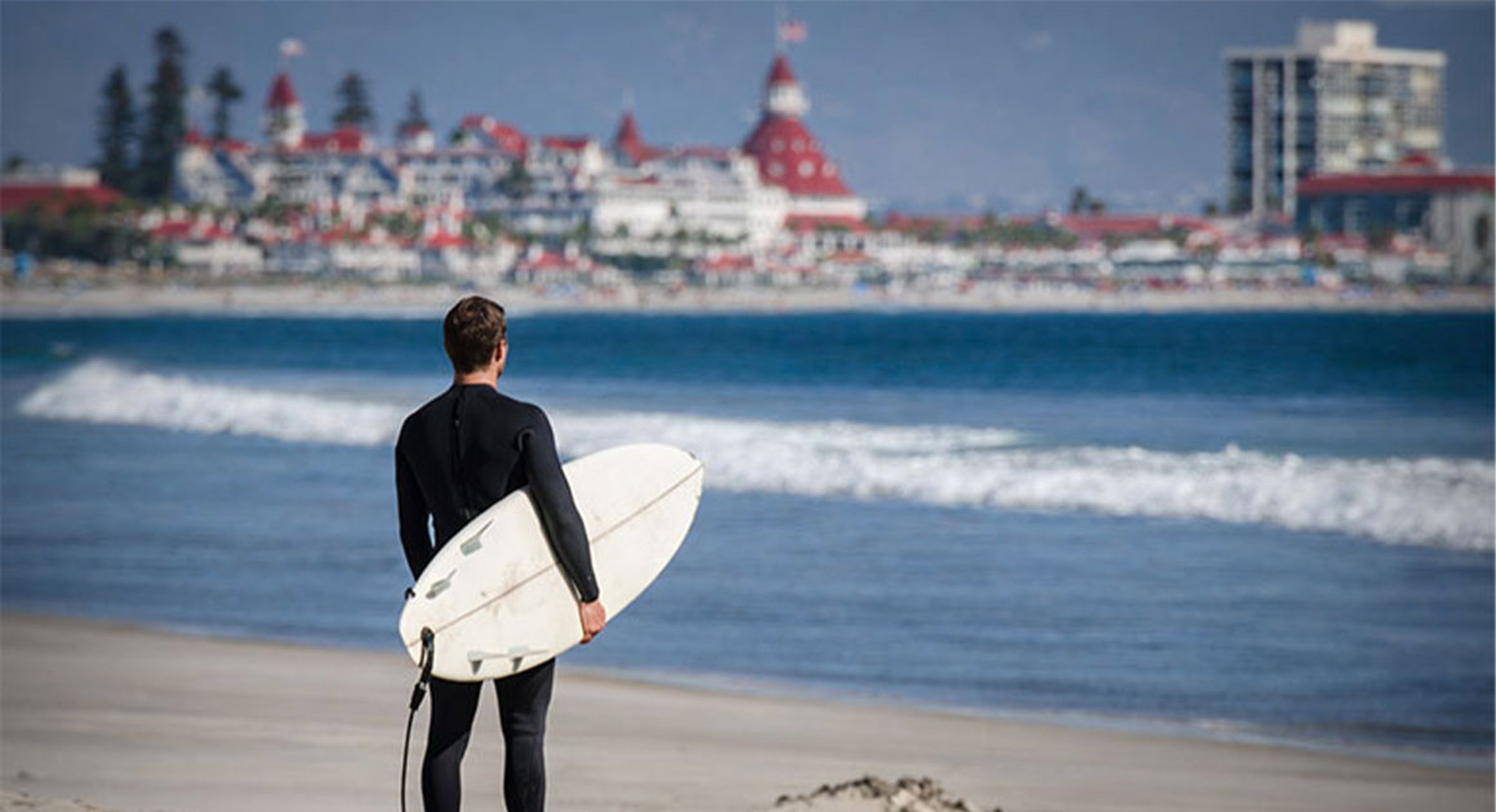 man holding surf board Hotel del Coronado in background