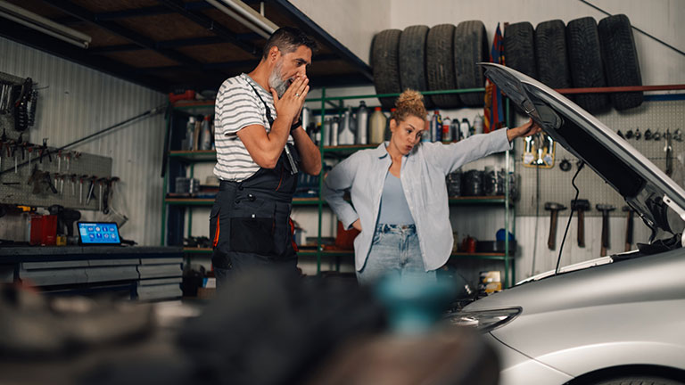 Shocked auto mechanic worker is looking under car hood with female customer. Side view of serviceman standing at auto mechanic workshop with female customer and looking at malfunction in shock. Credit:zamrznutitonovi