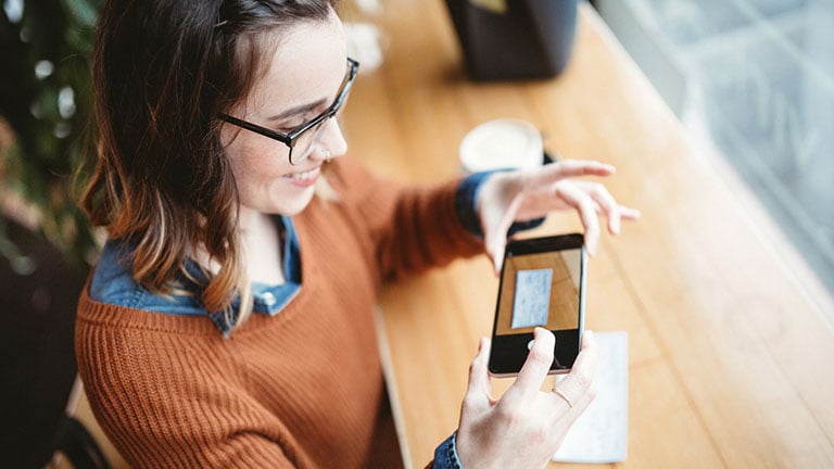 A smiling young woman takes a picture with her smart phone of a check or paycheck for digital electronic depositing, also known as "Remote Deposit Capture". She sits in a coffee shop, enjoying an espresso latte. Bright sunlight shines in the window. Horizontal image. Credit:RyanJLane