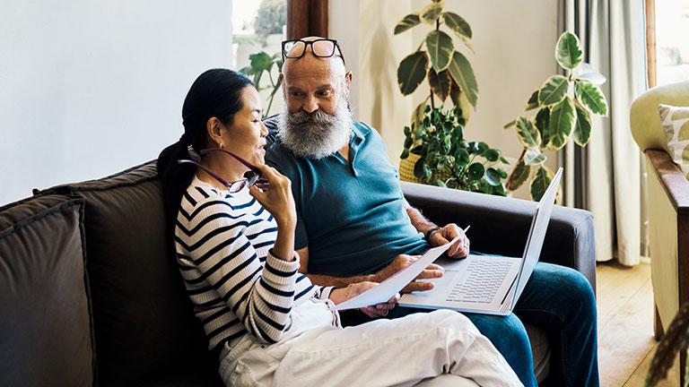 Senior interracial couple using a laptop and going through paperwork to pay bills online on the couch at home. Mature husband and wife talking and planning their retirement on a pc on the sofa Credit:pixdeluxe