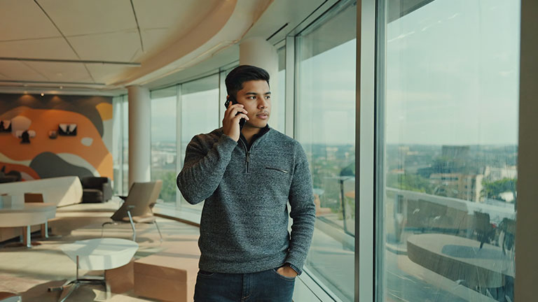 A indoor setting with a person standing near a window holding a smartphone to their ear. Their posture suggests attentiveness and caution. Casual, Background elements are soft and out of focus. No uniforms, no badges, no logos, no text. Clean, neutral corporate tone with cinematic realism.