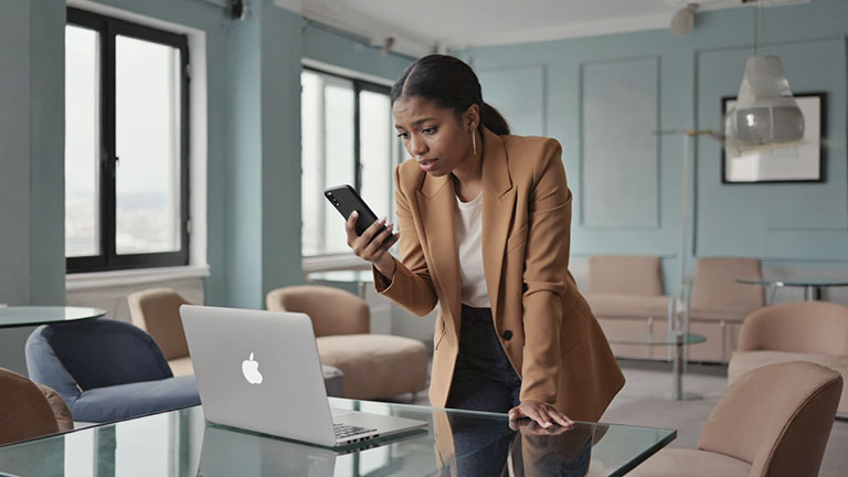 A realistic office-style setting showing a person holding a smartphone while a laptop sits open nearby. The person’s posture suggests hesitation or concern. Neutral tones, shallow depth of field, cinematic realism. Screens show generic interface shapes only—no text, no brand logos, no alerts.