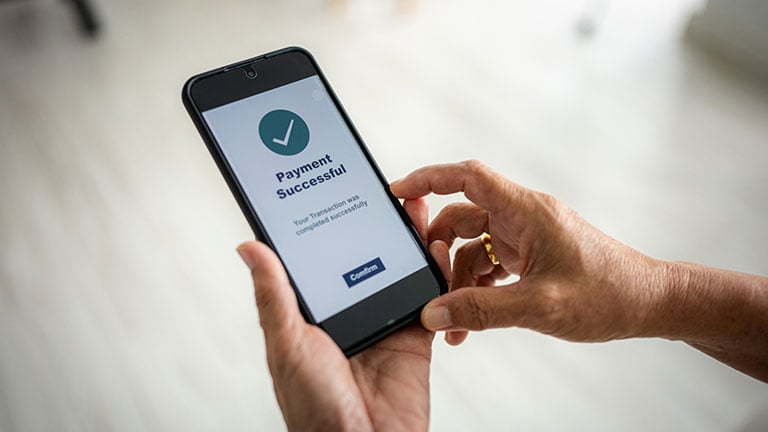 A senior woman is holding a smartphone displaying a payment successful message with a green check mark. Credit:simon2579