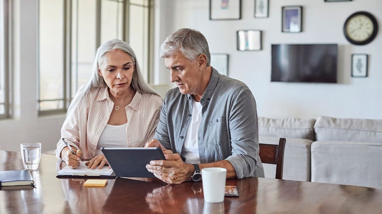 elder couple sitting at table looking at tablet and documents