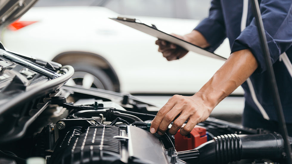 mechanic standing over open car hood with clipboard in hand