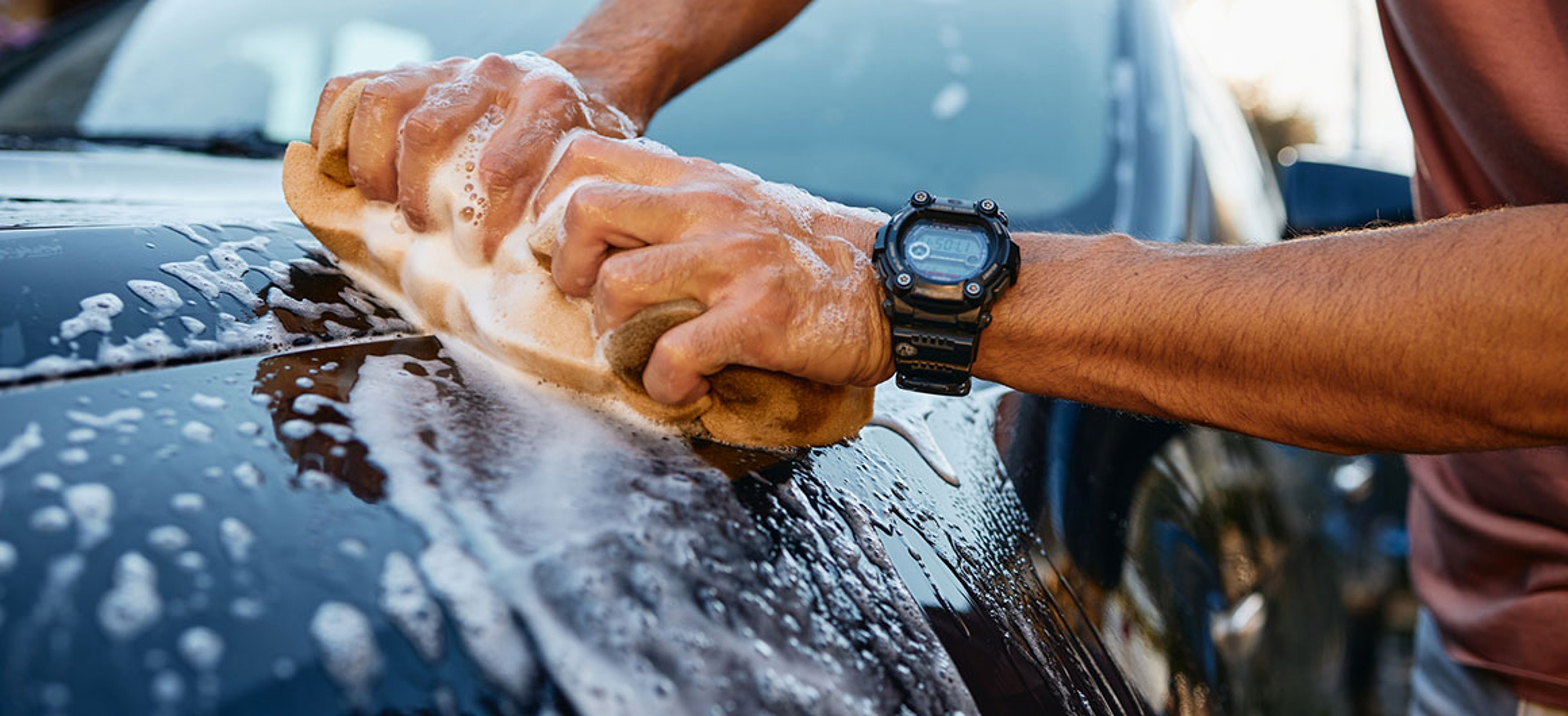 Close-up of a man washing a black car hood with a yellow sponge covered in soap suds outdoors during daytime. The man wears a black wristwatch and a brown shirt while cleaning the vehicle. Credit: SanyaSM