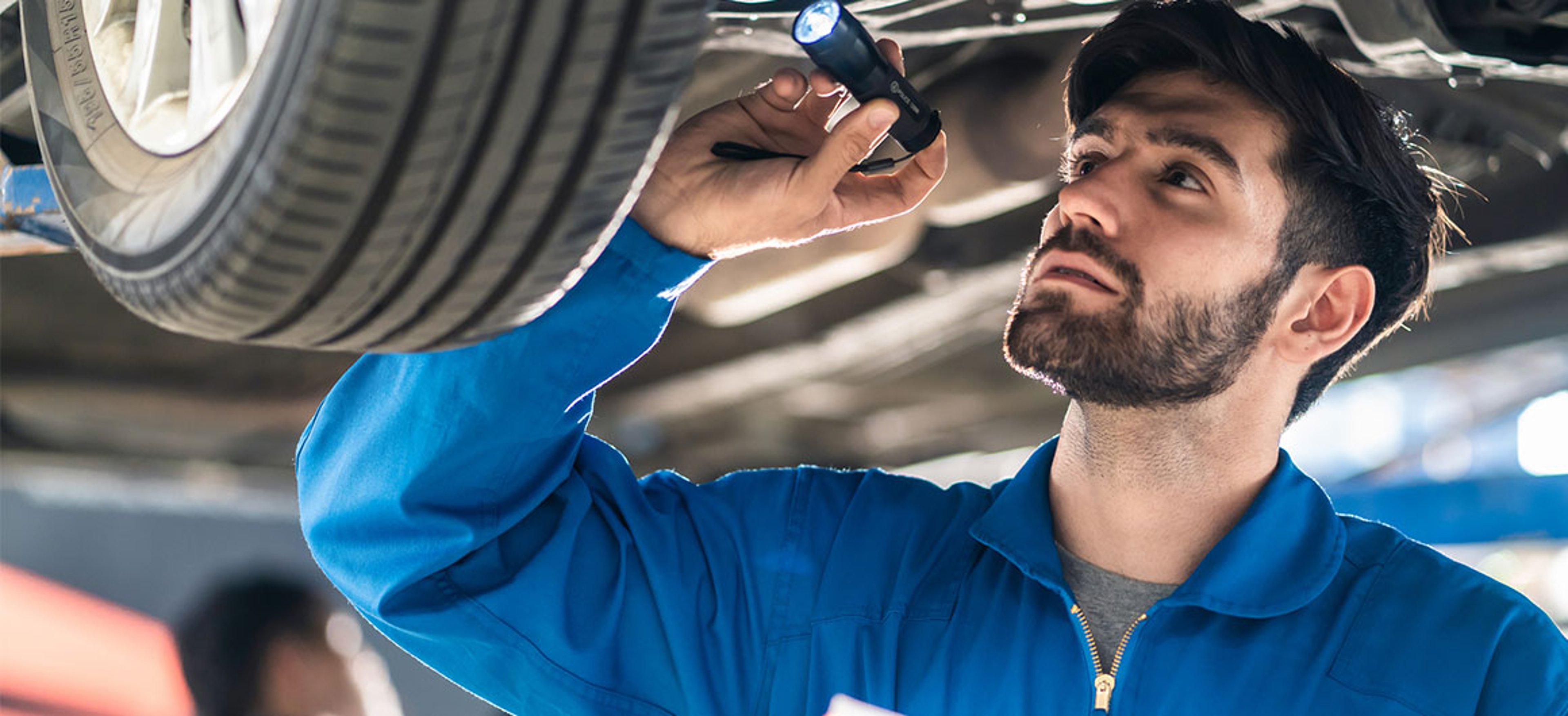 Vehicle service maintenance handsome man checking under car condition in garage. Automotive mechanic pointing flash light on wheel following maintenance checklist document. Car repair service concept stock photo by Kiwis