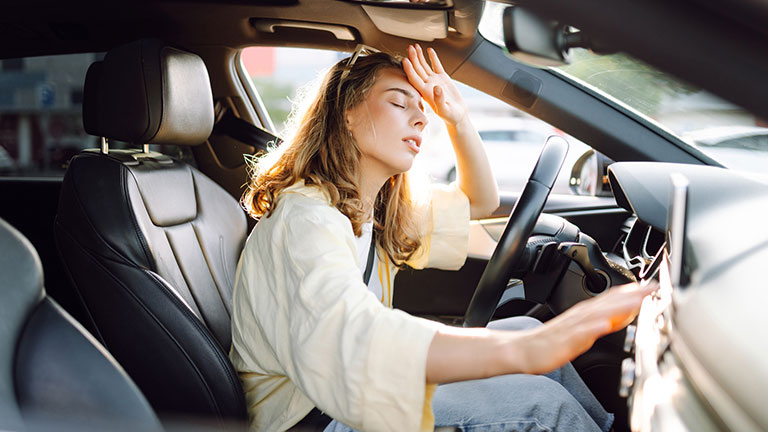 A young woman sits behind the wheel of a hot car. A female driver feels unwell due to the heat in the car on a sunny day. Concept of air conditioning, cooling, and transportation. Credit:Maksym Belchenko