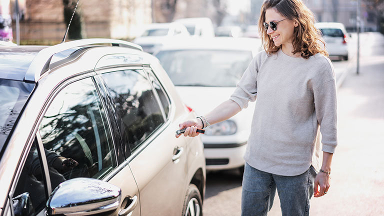 Happy cheerful young woman driver opens her car with a remote key on the street in the city; Credit:Olezzo