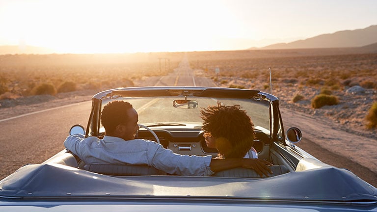 Rear View Of Couple On Road Trip Driving Classic Convertible Car Towards Sunset Credit: monkeybusinessimages