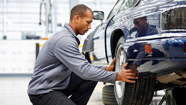 Full length of young engineer adjusting car tire while kneeling in showroom. Side view of technologist is repairing motor vehicle in industry. Male is manufacturing retro style automobile. Credit:Morsa Images