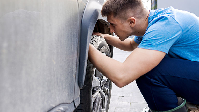 A man checks the condition of a car tire during maintenance on a driveway. The image portrays attention to detail, practical automotive care, and the importance of personal vehicle upkeep. Credit: vitapix 
