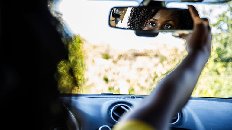 Over the shoulder view of young Black woman sitting in the driver's seat of her new car and adjusting the rear view mirror.