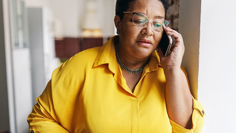 Serious african american senior woman in glasses and yellow shirt talking on phone with serious worried facial expression, receiving bad news from her doctor or relatives, leaning on wall Credit:shurkin_son