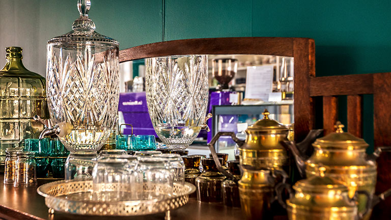 An antique drink decanter with glasses on a silver tray with brass serving ware setting in the foreground and colourful antique glassware in the background. Display is on top of an antique or vintage side table, console or side-board. Old bevelled mirror is in the background. Credit:marykan