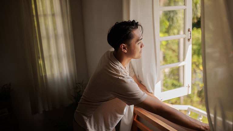 A smiling Asian man looks through the window after waking up in the morning. Credit: Johnce