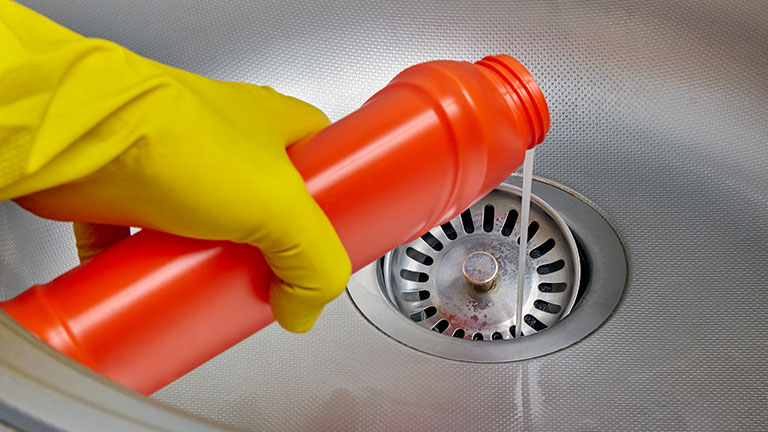 Person's hand in a yellow rubber glove pours pipe cleaner down the drain of a metal kitchen sink; Credit: Maksim Luzgin