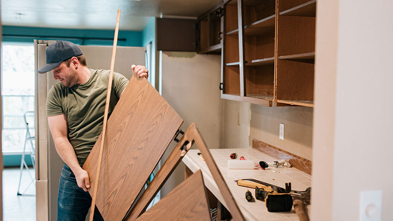 A Caucasian man does demolition work in the interior of his house for a renovation project. He pulls down old cabinets to make room for the new! Credit:RyanJLane