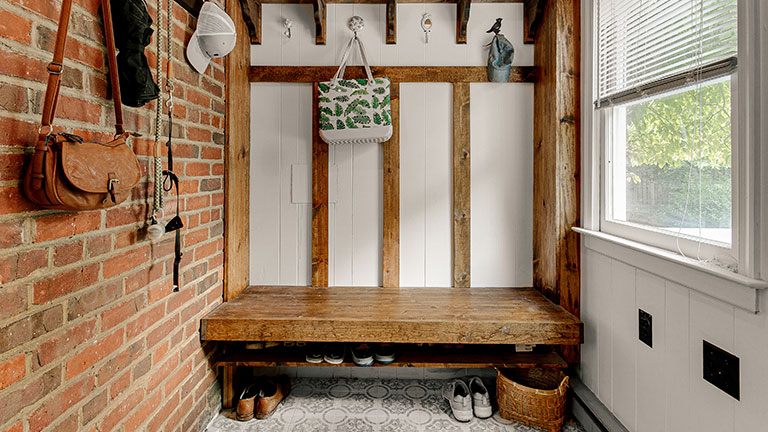 This image depicts a cozy and rustic mudroom with an exposed brick wall, a sturdy wooden bench, and multiple hooks holding an array of items, creating a practical entryway space with a warm and inviting atmosphere; Credit:Bryan Chavez