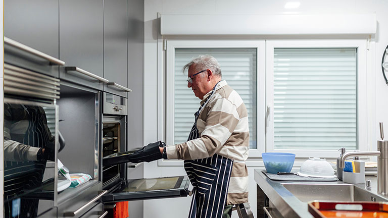 Senior chef taking a tray out of the oven using gloves and apron in a modern kitchen By Miguel Lifestyle