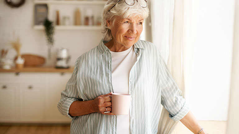 Coziness, domesticity and leisure concept. Portrait of stylish gray haired woman with round spectacles on her head enjoying morning coffee, holding mug, looking outside through window glass See Less By shurkin_son