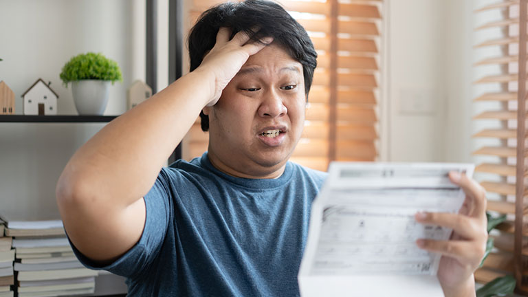 Asian sad and worried man going crazy with bill payment amount. Businessman worried with monthly bills to paid. Negative facial expressoion with paper overdue bill. Credit: StockerThings
