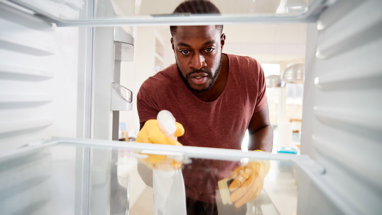 View Looking Out From Inside Empty Refrigerator As Man Wearing Rubber Gloves Cleans Shelves By Monkey Business