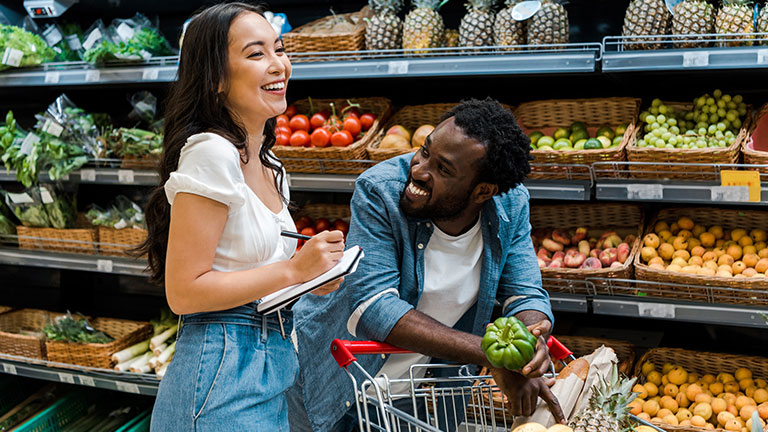 happy african american man looking at asian woman with notebook near shopping cart with groceries  by LIGHTFIELD STUDIOS