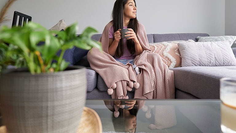 a female sitting down comfortably on a sofa smiling, with a cozy blanket wrapped around her, drinking coffee Credit:ali Quevedo