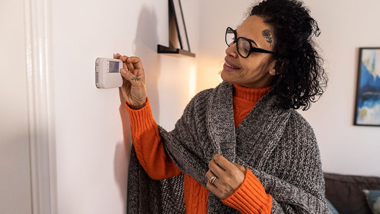 A shot of a multicultural female homeowner changing the temperature of her central heating thermostat in her house. She is wearing a blanket around her shoulders. Credit:SolStock
