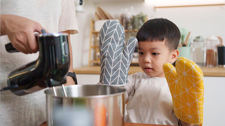 Young Asian boy learning to cook with his mother in a cozy kitchen, wearing colorful patterned oven mitts, observing the preparation process with curiosity and focus; by eggeeggjiew