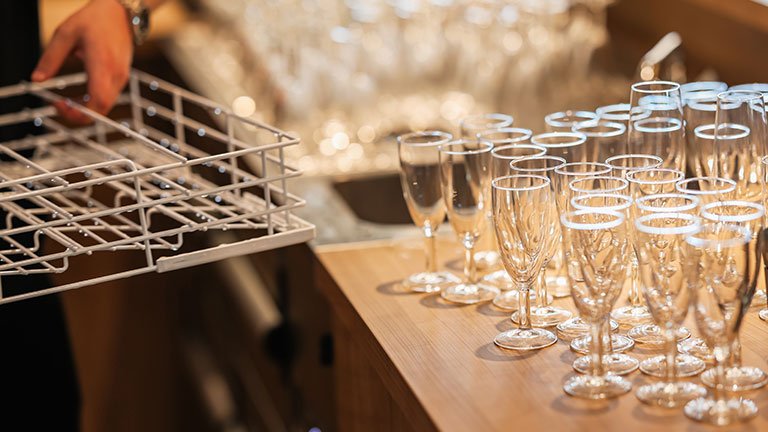 Clean drinking glasses arranged on counter next to a dishwasher rack in a restaurant kitchen. Preparation scene before service, no recognizable people; by Samir