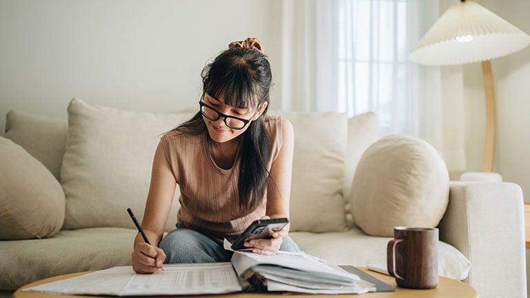A woman sits at her living room with smartphone and financial reports doing her monthly budget. Credit: staticnak1983
