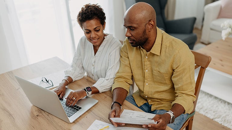 A focused couple is seen working on their budget plans, with one using a laptop and the other reviewing documents on the table. They show determination and cooperation. Credit: supersizer