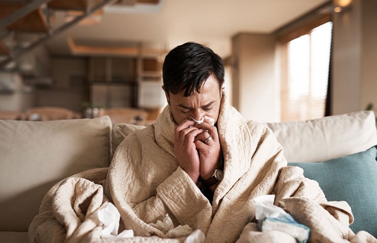 man wrapped in blanket, on couch, holding tissue to nose
