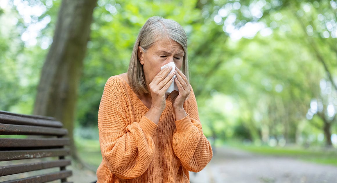 woman sneezing into tissue while sitting on park bench
