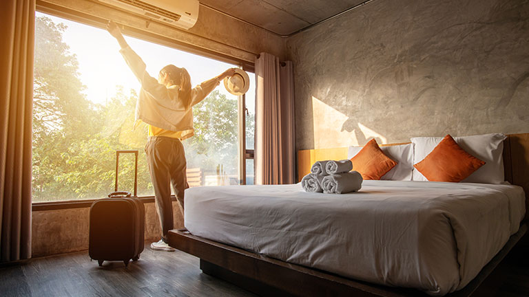 Portrait of tourist woman raised her hands and standing nearly window, looking to beautiful view with her luggage in hotel bedroom after check-in. Credit: Boyloso