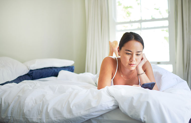 girl laying on white bedding in naturally lit room on her phone
