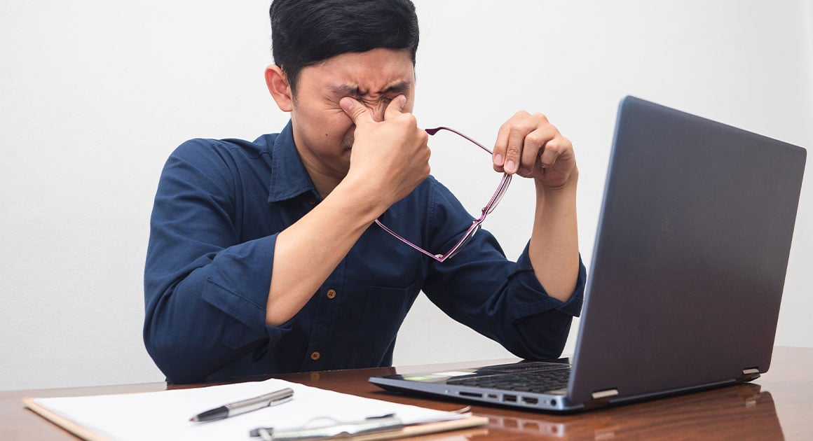 man rubbing eyes, holding glasses, laptop on desk in front of him