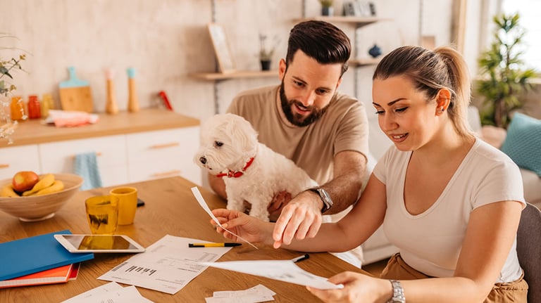 Young heterosexual couple sitting in their living room and checking their finances. Man holding cute toy dog in his lap. Credit: urbazon