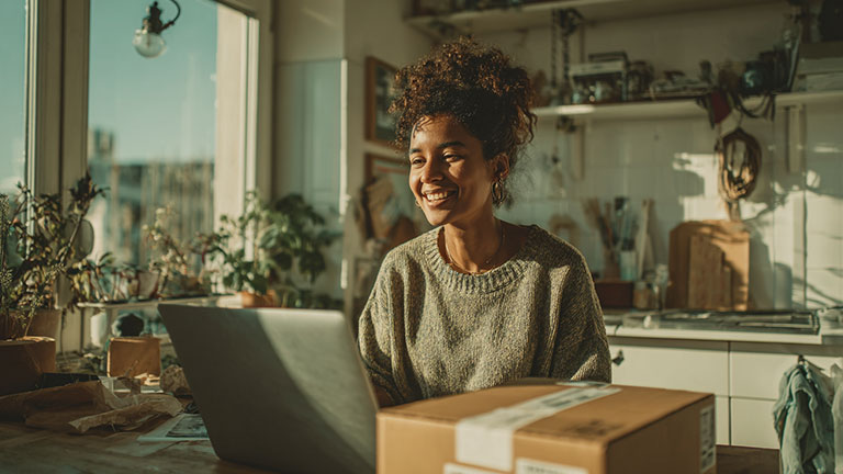 Bright editorial image of a smiling woman in her 30s at a kitchen counter, ready to ship a package, laptop open with online store visible. Cardboard box, tape, and label in front of her. Sunlight through window, warm modern colors (soft white, beige, teal). Clean, minimal layout with space for headline text on left. Commercial lifestyle photo, optimistic tone.