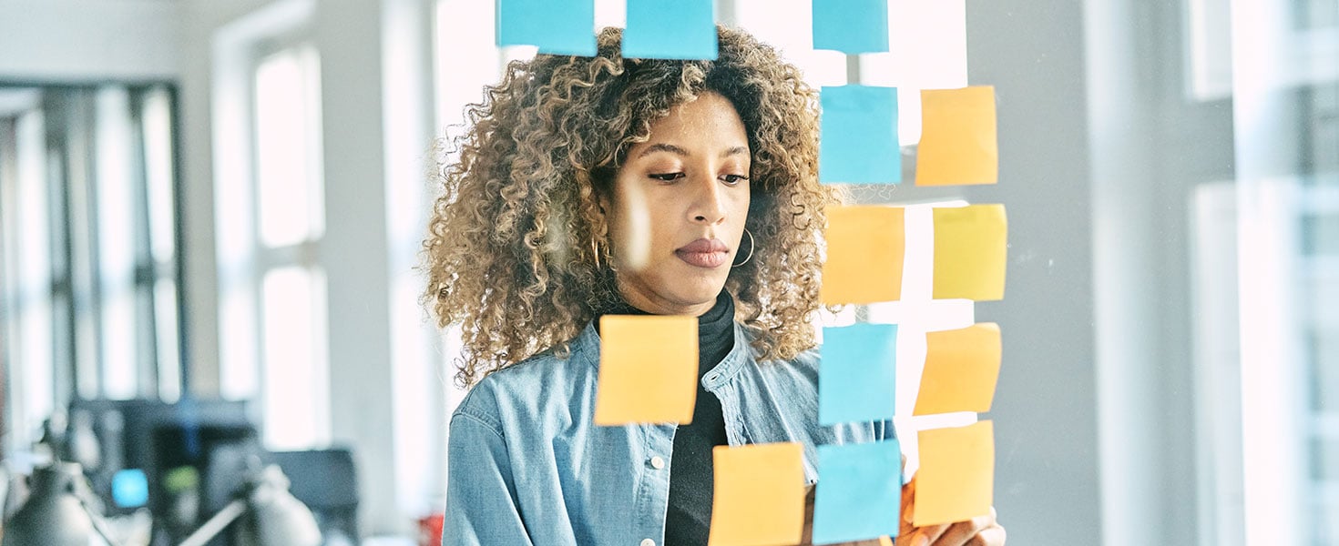 Young woman putting sticky notes on a glass wall while working on a new project in a startup office, engaged in strategic planning and creative thinking. Credit:Portra