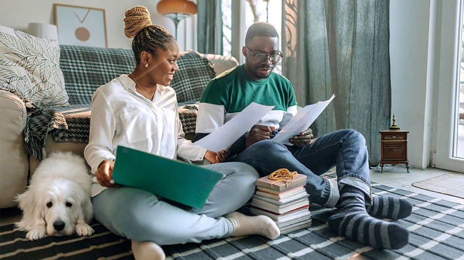 man and woman looking at papers while sitting on floor next to dog and a pile of books with a chain on top of them
