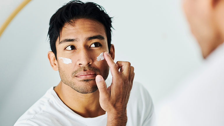 Man applying cream, lotion and moisturizer for a skincare routine while grooming in a mirror at home. Handsome young guy using sunscreen lotion with spf for uv protection on his face for healthy skin; credit:Delmaine Donson